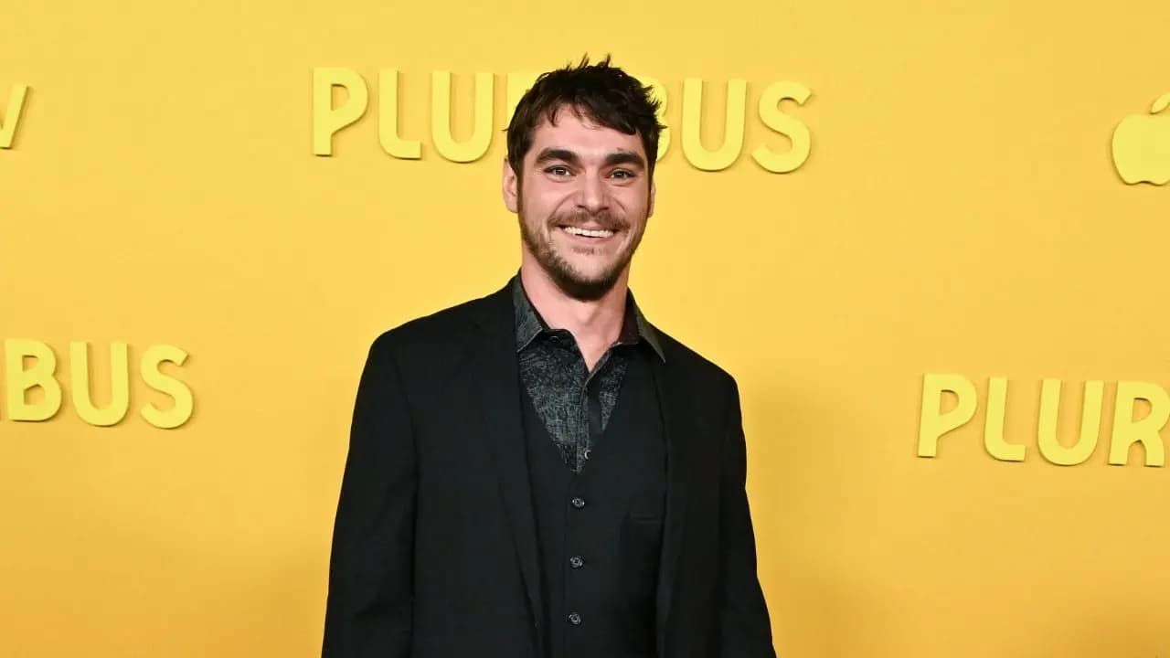 A black-and-white photo of RJ Mitte smiling, holding a clipboard, wearing a blue jacket. Background blurred, like a memory.