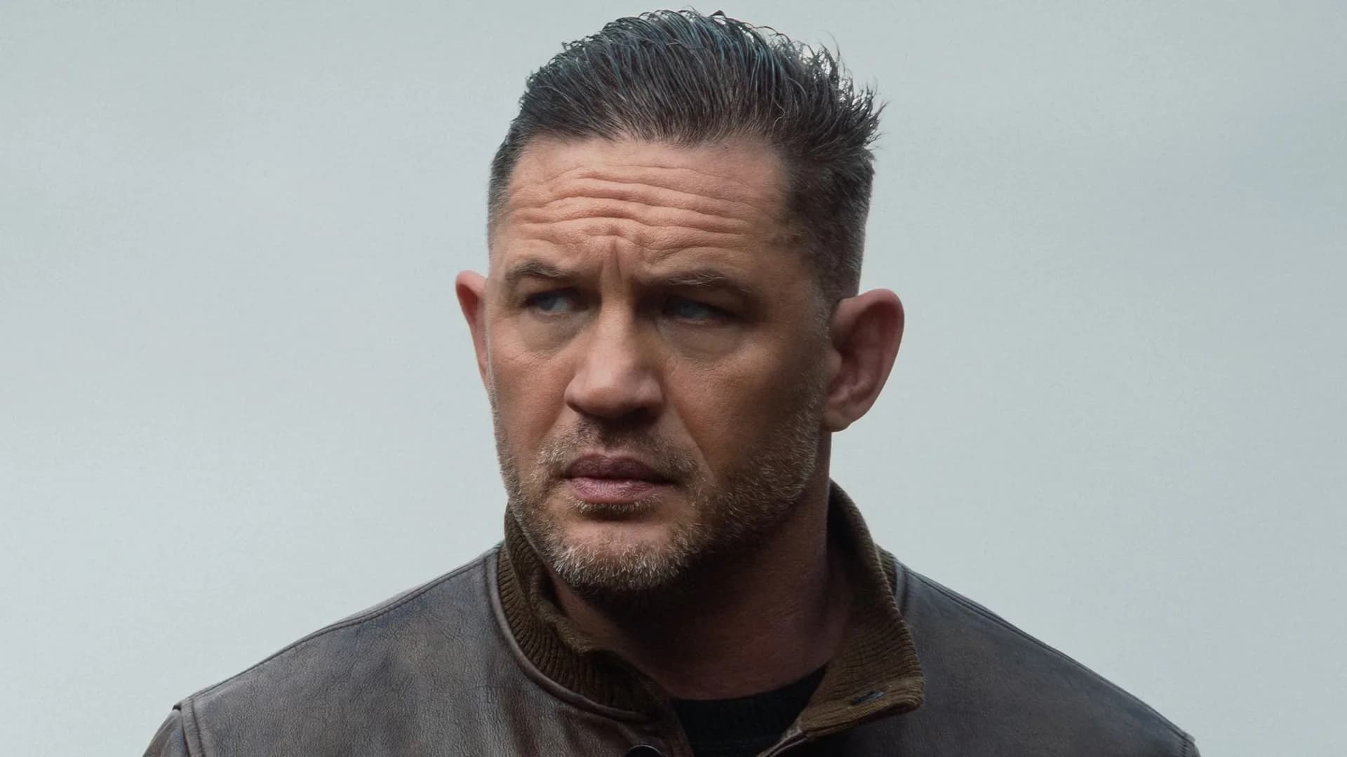Tom Hardy in character as James Delaney from Taboo, standing in a dimly lit room with a cigarette in hand.