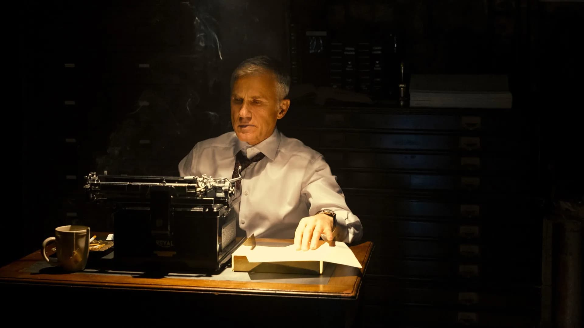 Christoph Waltz in a dark suit, standing in a dimly lit office corridor, staring ahead with unnerving stillness.
