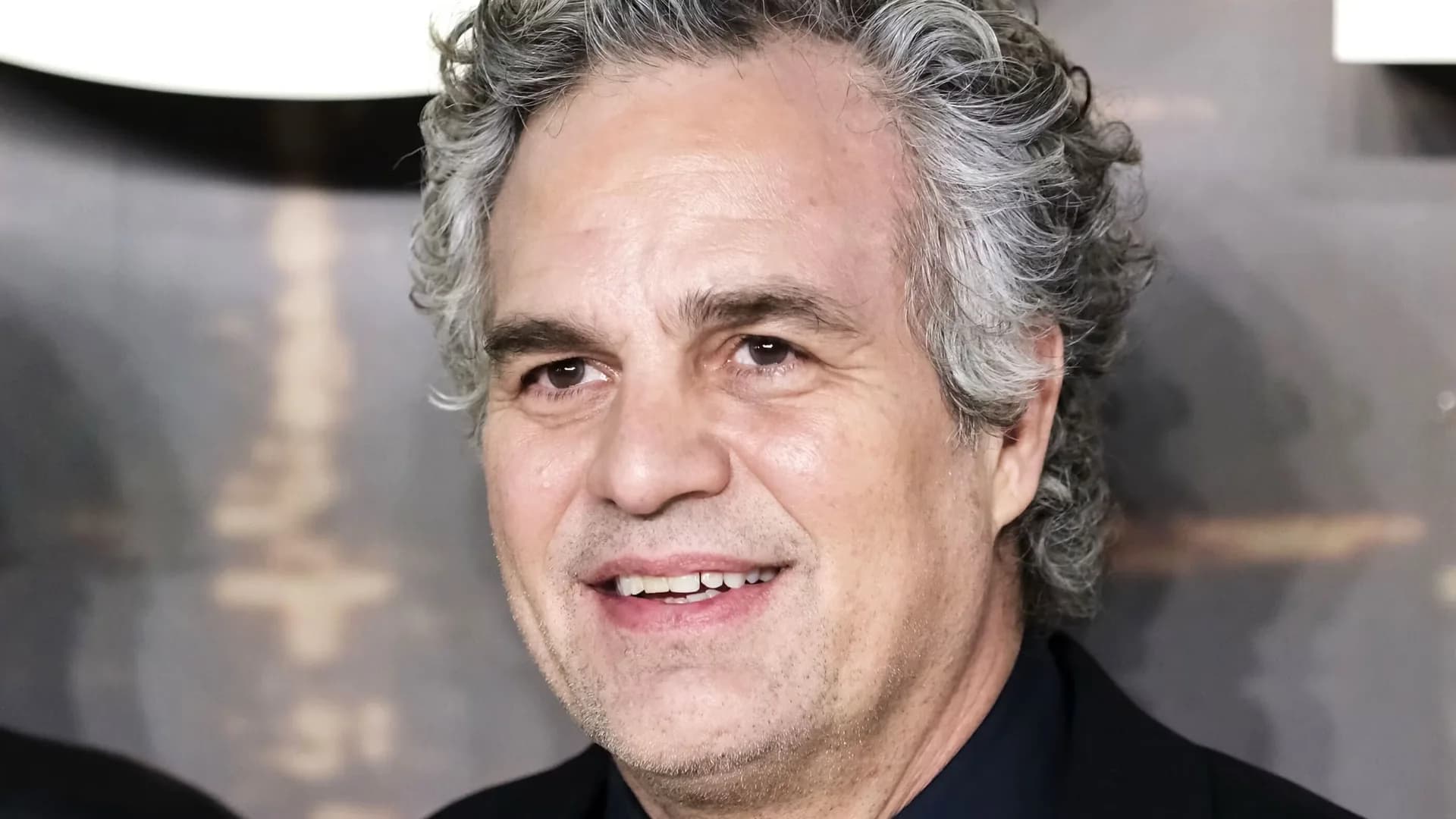 Mark Ruffalo in a sleek black suit, standing in front of a mirrored wall, looking intense.
