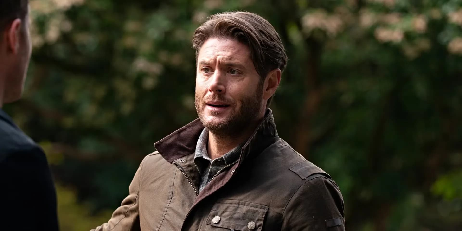 Jensen Ackles in a dark, intense scene, standing in a dimly lit urban setting, looking determined.