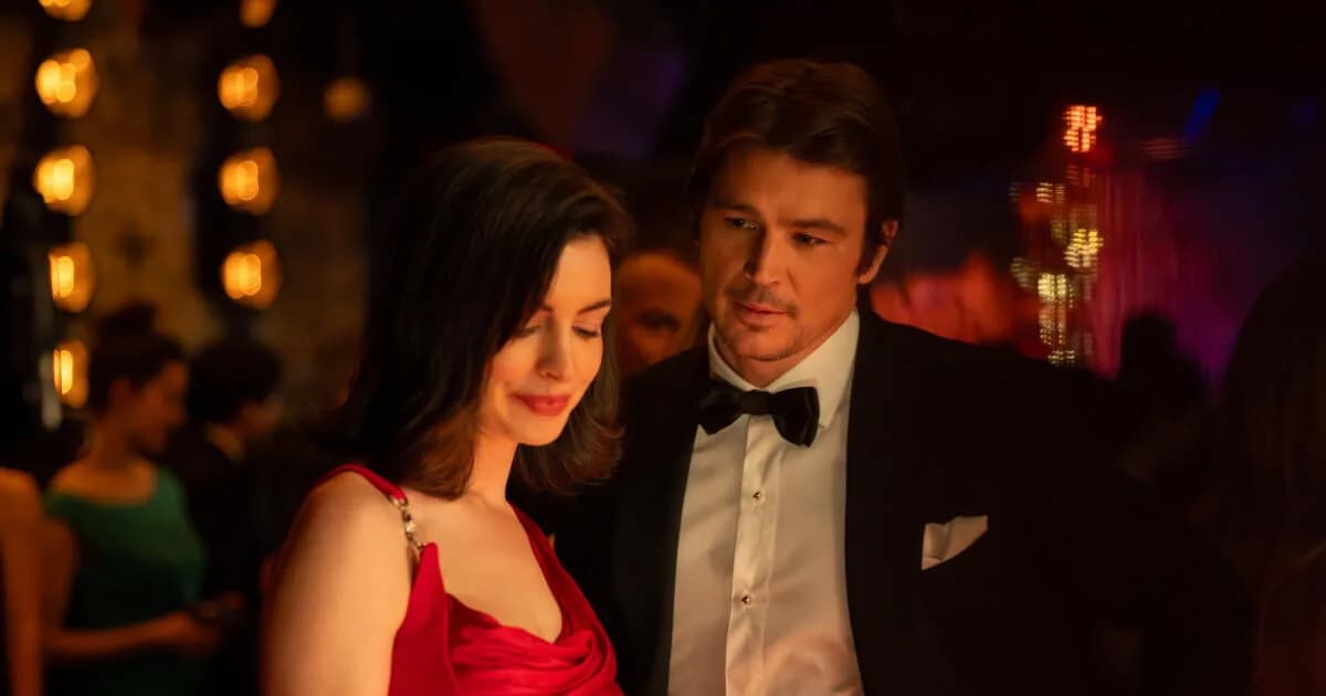 Anne Hathaway in red, eyes closed, seated in a dimly lit room. A single light casts shadows across her face.