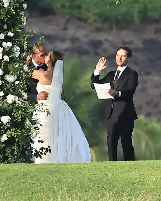 Tobey Maguire stands at a Hawaiian beachside ceremony, reading from a piece of paper, smiling as Jennifer Meyer and Geoff Ogunlesi exchange vows.