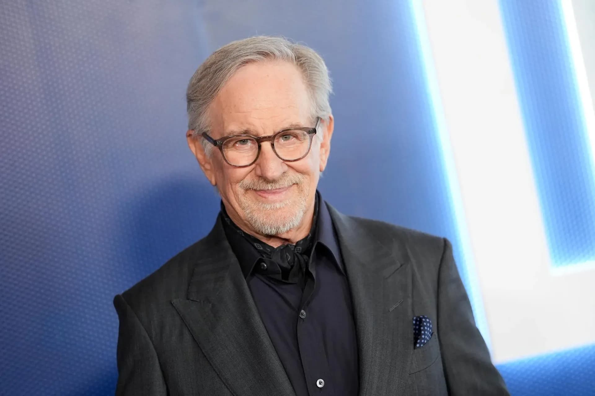 Steven Spielberg in a suit, standing in front of a giant Transformer sculpture, looking thoughtful.