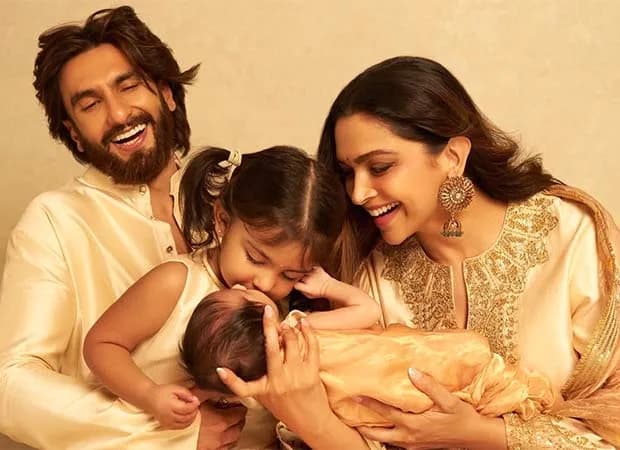 Deepika Padukone holding her newborn daughter Devi, with her older daughter Dua kissing the baby’s forehead, and Ranveer Singh standing beside them in an ivory kurta.
