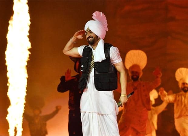 Diljit Dosanjh performing on stage during a stadium concert, wearing a colourful outfit and turban, with a large crowd visible under stage lighting.