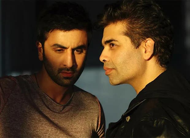 Karan Johar smiling at an event, with a still from Ae Dil Hai Mushkil featuring Ranbir Kapoor and Anushka Sharma in the background